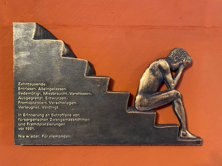 Copper bas-relief on an orange background. On the lowest step of a staircase, a child sits, hands resting on knees, and head buried in palms. The inscription on the memorial plaque reads: Tens of thousands, torn away, abandoned, humiliated, abused, rejected, excluded, uprooted, placed in foster care, silenced, denied, indentured. In memory of those affected by welfare coercive measures and foster placements before 1981. Never again. For no one.