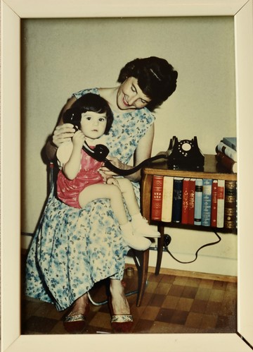 Jasmin Schweizer*, who is around 2 years old, is sitting on her mother's lap and holding a large black telephone receiver in her hand. Her mother leans towards her and smiles. A black telephone with a white dialling disc stands on a small bookcase to her right.