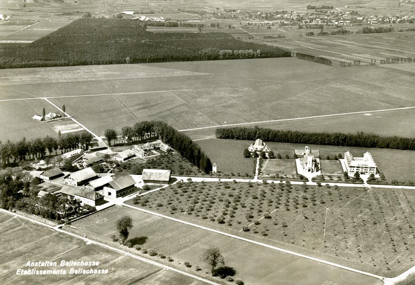 The aerial view shows the internees' barracks on the left, as well as the workshops and farm buildings, surrounded by trees. On the right are the director's villa, the church and the prison wing.