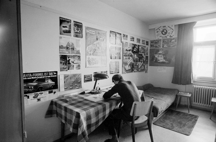 Black and white photo of a room in the Uitikon-Waldegg “correctional labour facility”. A teenager is sitting at his desk, to the left of which is a bed with a carpet in front of it. Curtains hang at the window, and posters of sports cars adorn the walls.