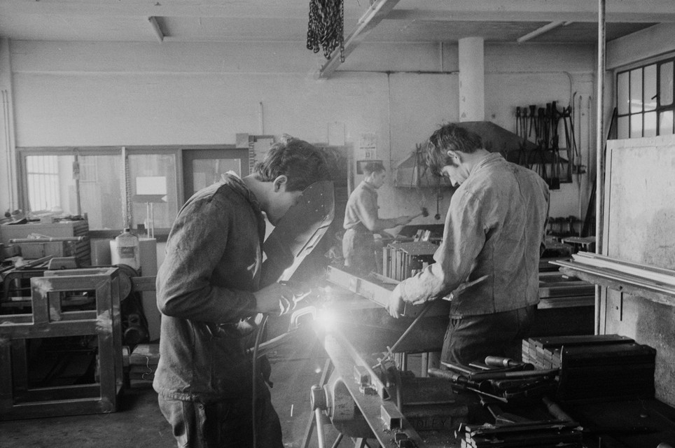 Three apprentices are working in a locksmith's workshop. In the foreground, welding is taking place, while further back, metal is being worked on with tools